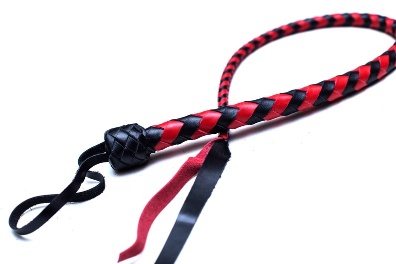 Op de afbeelding: Een zwarte en rode leren zweep met een gevlochten ontwerp. De zweep heeft een zwart leren handvat en een rood-zwarte gevlochten riem.