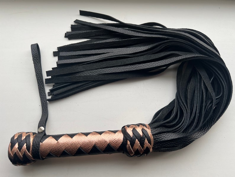 May include: A black leather flogger with a braided handle featuring copper-colored accents. The handle has a loop for hanging. The flogger's strands are long and supple, and the overall design is sleek and functional.