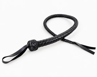 Könnte beinhalten: Eine schwarze Lederpeitsche mit geflochtener Struktur, einem runden Knoten am Griffende und einer Handschlaufe. Die lange, flexible Schnur ist zu einer Schlaufe gewunden und endet in zwei flachen, dünnen Lederstreifen am Ende des geflochtenen Teils.