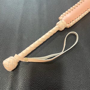 May include: A light brown leather whip with a braided handle and a loop at the end. The whip is on a dark brown textured surface.