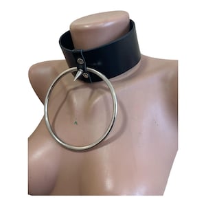 May include: Black leather choker with a large silver ring and a single silver spike. The choker is fastened with two silver rivets. The accessory is displayed on a mannequin.