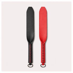 May include: Two leather paddles are shown, one black and one red. Each paddle has a long, slender form with a rounded end and a stitched handle. A black metal ring is attached to the base of each handle.