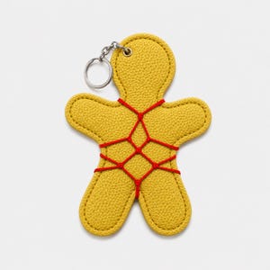 Op de afbeelding: Een mosterdgele, getextureerde sleutelhanger in de vorm van een peperkoekmannetje van leerachtig materiaal. Het figuurtje is versierd met een rood koord dat in een kruispatroon over de romp en ledematen is geregen, inclusief een zilveren ring.