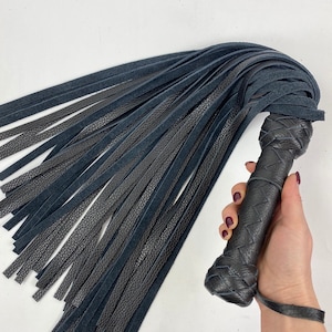 May include: A black leather flogger with a braided handle and numerous long, thin leather falls. The handle is intricately woven, and a wrist strap is attached. The falls are a mix of smooth and textured leather.