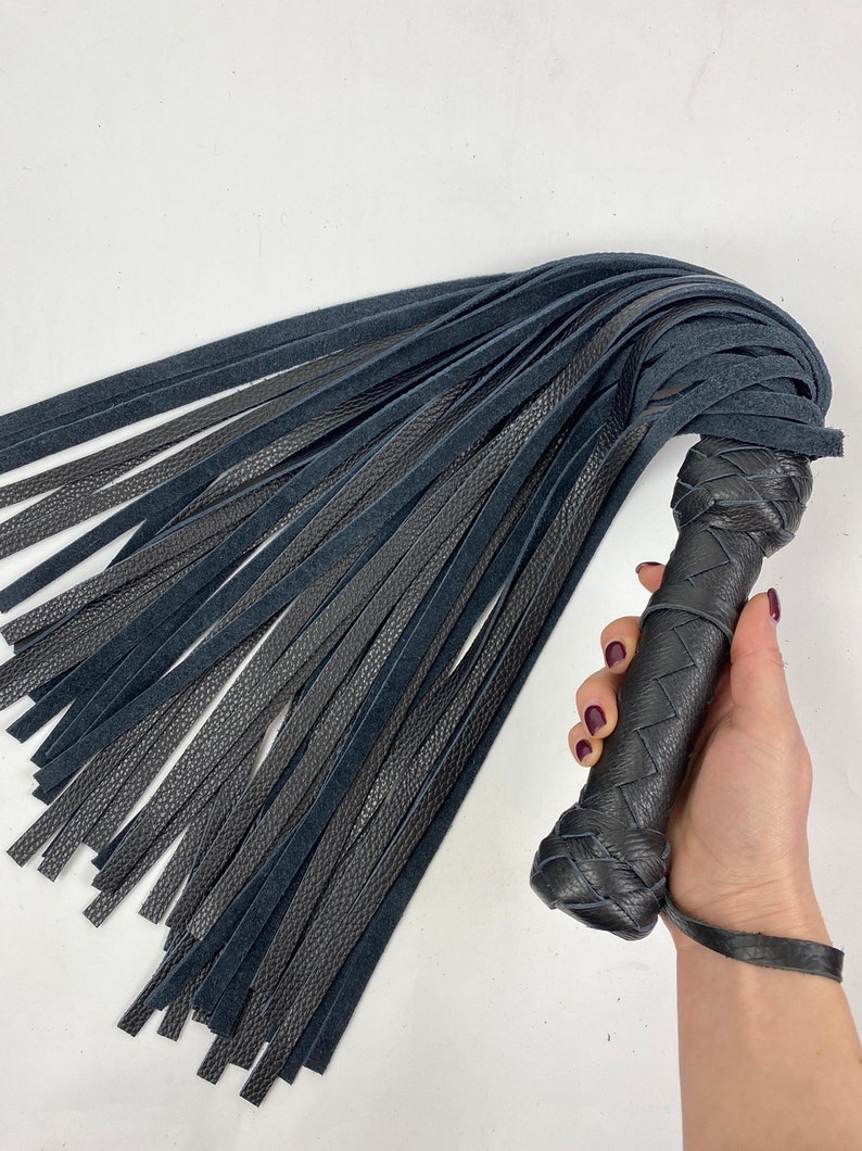 May include: A black leather flogger with a braided handle and numerous long, thin leather falls. The handle is intricately woven, and a wrist strap is attached. The falls are a mix of smooth and textured leather.