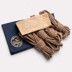Op de afbeelding: Een verzameling bruine touwbundels, een marineblauwe gevouwen doek met een gouden embleem en een beige doek met kleurrijke spikkels. Het touw is strak gewikkeld. De items zijn op een wit oppervlak geplaatst.