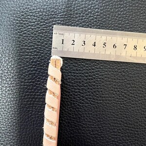 May include: A close-up of a pink leather cord with a braided design. The cord is measured with a ruler, showing it is approximately 3.5 inches long.