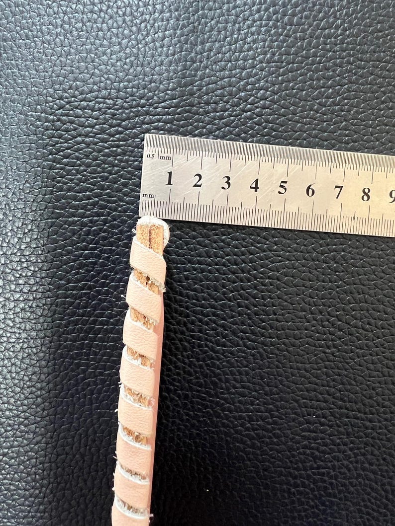 May include: A close-up of a pink leather cord with a braided design. The cord is measured with a ruler, showing it is approximately 3.5 inches long.