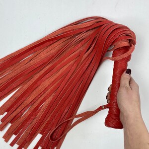 May include: A red leather flogger with a braided handle and numerous long, thin falls. The handle features a wrist strap. The flogger is held against a white background.