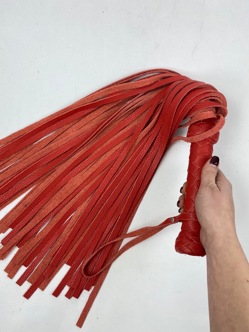 May include: A red leather flogger with a braided handle and numerous long, thin falls. The handle features a wrist strap. The flogger is held against a white background.