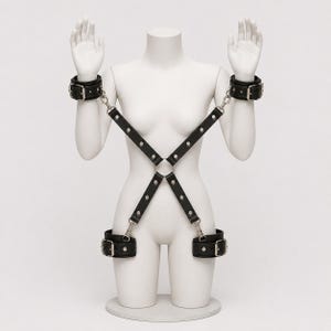 May include: A black leather harness featuring adjustable wrist and ankle cuffs connected by silver-toned metal hardware. The straps form an X-shape across the torso, secured with a central metal ring and metal buckles on each of the four cuffs.