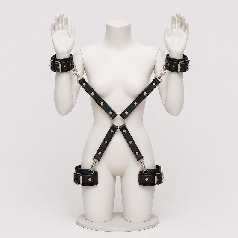 May include: A black leather harness featuring adjustable wrist and ankle cuffs connected by silver-toned metal hardware. The straps form an X-shape across the torso, secured with a central metal ring and metal buckles on each of the four cuffs.