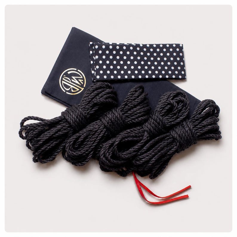 May include: Four coils of black rope arranged in a row, accompanied by a dark navy blue fabric pouch featuring a gold circular logo and a separate rectangular fabric piece with a white polka dot pattern on a dark navy blue background.