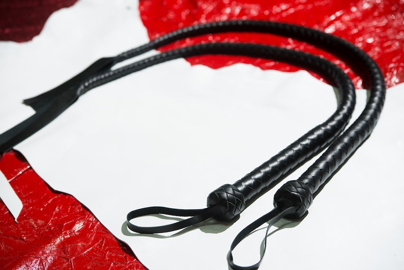Op de afbeelding: Twee zwarte leren zweepen met gevlochten handvatten en lussen. De zweepen liggen op een wit oppervlak met een rode achtergrond.
