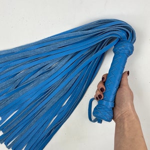 May include: A blue leather flogger with a braided handle and numerous long, thin leather falls. The handle has a loop for grip. The flogger is a solid blue color.