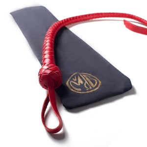 Op de afbeelding: Een rode leren zweep met een gevlochten handvat en een lus aan het einde. De zweep ligt op een donkerblauwe stoffen tas met een gouden monogram.