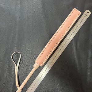 May include: A light pink leather paddle with a braided edge and a knotted handle. The paddle is 50 cm long and has a ruler printed on one side.