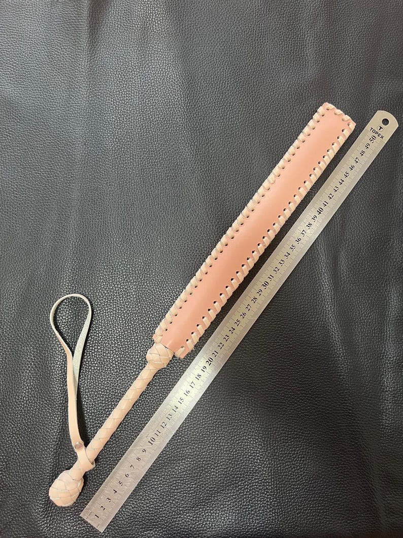 May include: A light pink leather paddle with a braided edge and a knotted handle. The paddle is 50 cm long and has a ruler printed on one side.