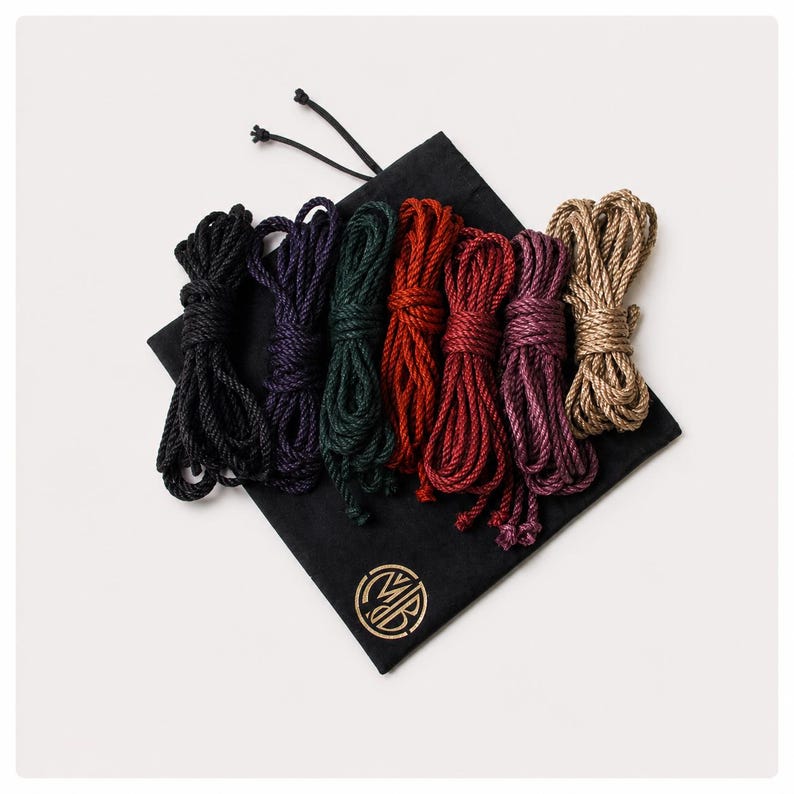 May include: A black drawstring storage bag featuring a gold circular logo with the letters MB, topped with seven coiled lengths of rope in black, dark purple, dark green, bright red, dark red, magenta, and tan, arranged in a horizontal row.