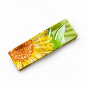 May include: A rectangular fabric headband featuring a large yellow sunflower graphic with a brown center. The background consists of vibrant green leaves and a white diagonal striped pattern on the right side, creating a bold floral design.
