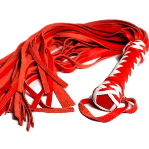 May include: A red leather flogger with a white and red handle. The handle is wrapped in a zig-zag pattern. The flogger's strands are long and thin, and the handle has a loop at the end.