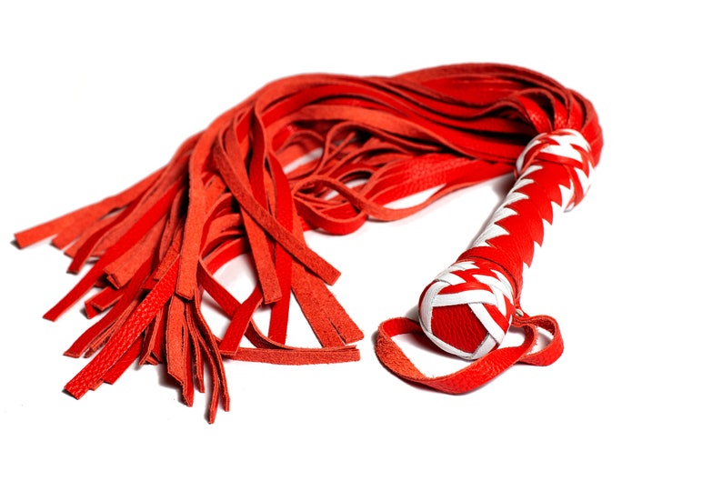 May include: A red leather flogger with a white and red handle. The handle is wrapped in a zig-zag pattern. The flogger's strands are long and thin, and the handle has a loop at the end.