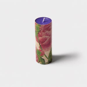 May include: A cylindrical pillar candle featuring a vibrant floral print of pink peonies with green stems and leaves against a cream background. The top surface of the candle is a solid deep purple color with a single white cotton wick centered.