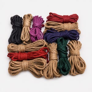 Può includere: Una collezione di dodici fasci di corda intrecciata disposti a griglia. Le corde sono disponibili in vari colori solidi, tra cui nero, beige, viola, rosso scuro, blu navy e verde bosco, ciascuna ordinatamente avvolta e legata al centro.
