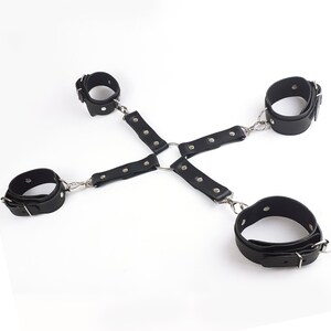 May include: A black four-point restraint harness featuring four adjustable cuffs connected by leather straps to a central metal O-ring. The straps are secured with silver-toned metal rivets and buckles, creating a symmetrical cross-shaped configuration.