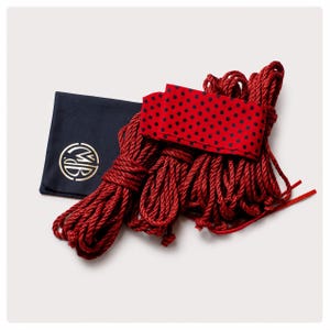 May include: A set of deep red twisted ropes bundled together, accompanied by a red fabric strip featuring a black polka dot pattern and a dark navy blue square pouch displaying a circular gold logo with the letters MB in the center.