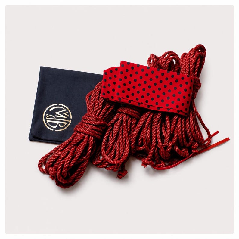 May include: A set of deep red twisted ropes bundled together, accompanied by a red fabric strip featuring a black polka dot pattern and a dark navy blue square pouch displaying a circular gold logo with the letters MB in the center.