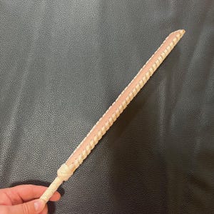 May include: A long, thin, braided leather whip with a light brown color and a pink undertone.