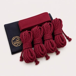 May include: Four coiled lengths of deep red rope arranged in a row, accompanied by a folded deep red fabric band and a dark navy blue rectangular fabric pouch featuring a circular gold logo with the letters MB printed on the front surface.