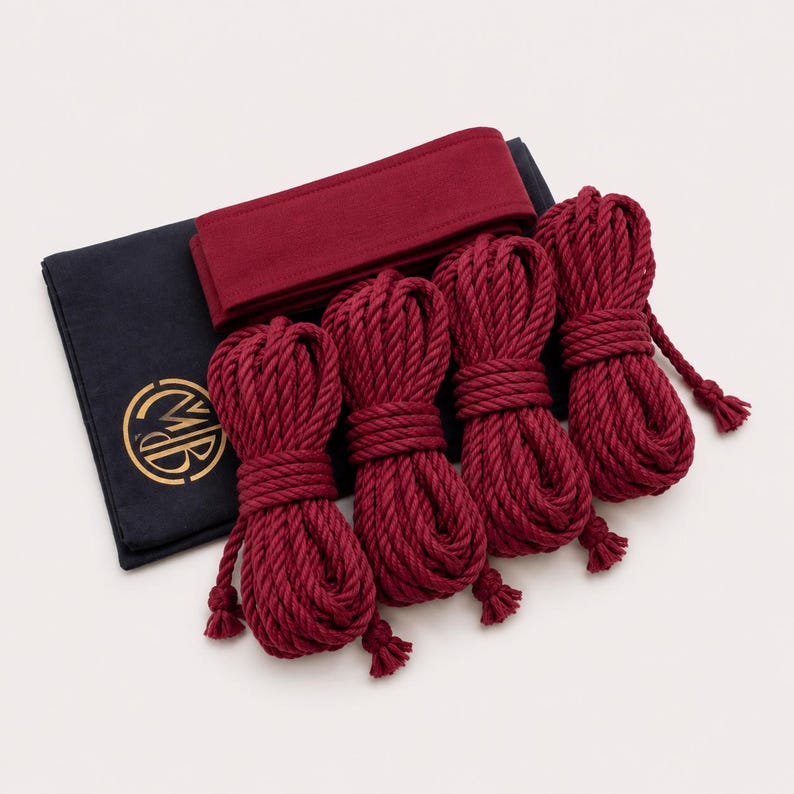 May include: Four coiled lengths of deep red rope arranged in a row, accompanied by a folded deep red fabric band and a dark navy blue rectangular fabric pouch featuring a circular gold logo with the letters MB printed on the front surface.