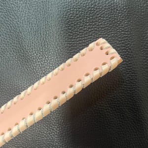 May include: A pink leather strap with a white stitched edge. The strap is on a black textured surface.