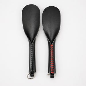 May include: Two black leather paddles with oval heads and textured handles. One paddle has a silver ring at the base, the other has red stitching along the handle and a silver ring.