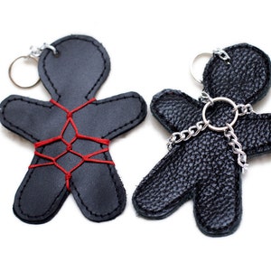 Op de afbeelding: Twee zwarte leren sleutelhangers in de vorm van mensen. De ene sleutelhanger is vastgebonden met rood touw, de andere met een zilveren ketting.