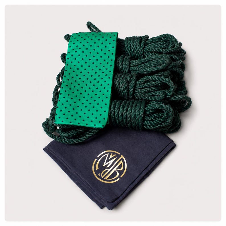 May include: A set of dark green coiled ropes bundled together with a bright green fabric wrap featuring a black polka dot pattern. Below the ropes lies a folded dark navy blue cloth square with a gold circular logo containing the letters MB.