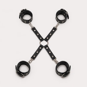 May include: A black leather restraint harness featuring four adjustable cuffs connected by straps to a central metal ring. The straps are secured with silver-toned rivets and metal swivel clips, forming an X-shaped configuration for limb positioning.