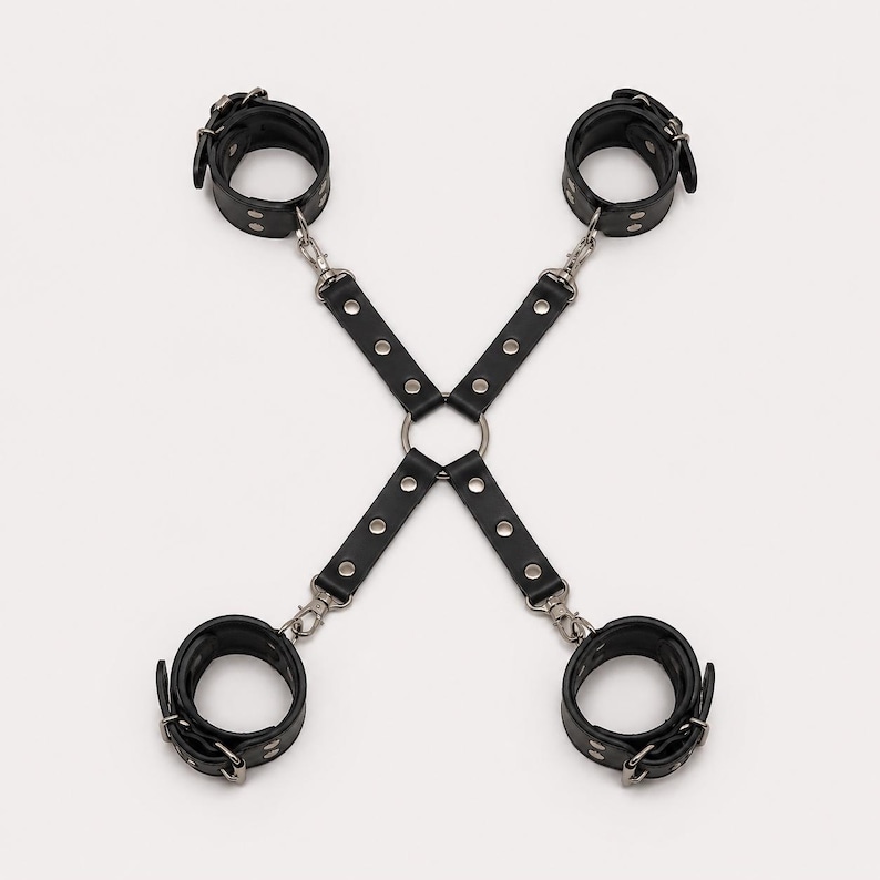 May include: A black leather restraint harness featuring four adjustable cuffs connected by straps to a central metal ring. The straps are secured with silver-toned rivets and metal swivel clips, forming an X-shaped configuration for limb positioning.
