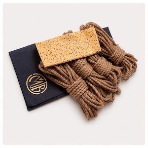 May include: A dark navy blue fabric pouch featuring a gold circular logo with the letters MB, accompanied by several coils of thick brown jute rope and a rectangular yellow fabric strip decorated with a colorful multi-colored confetti pattern.