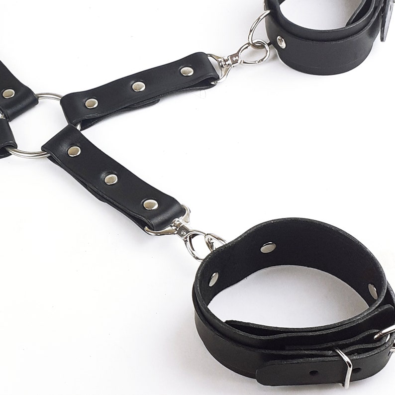 May include: A black leather harness featuring two adjustable wrist cuffs connected by silver-toned metal rings, rivets, and swivel lobster clasps. The structure consists of multiple leather straps joined at a central ring, creating a modular design.