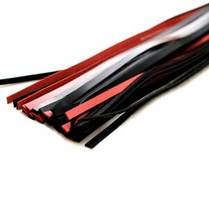 Puede incluir: Primer plano de un l&aacute;tigo de cuero negro con hebras de cuero rojo y gris. El l&aacute;tigo est&aacute; sobre una superficie blanca.