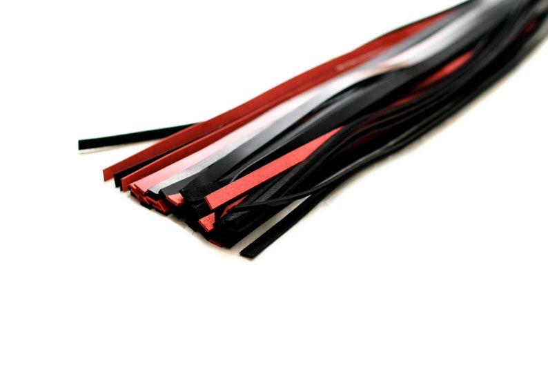 Puede incluir: Primer plano de un l&aacute;tigo de cuero negro con hebras de cuero rojo y gris. El l&aacute;tigo est&aacute; sobre una superficie blanca.