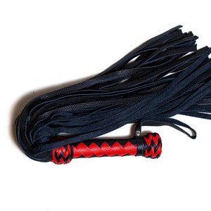May include: A black leather flogger with a red and black handle. The flogger features multiple black leather falls and a handle with a red center and black braided ends. The item is set against a white background.