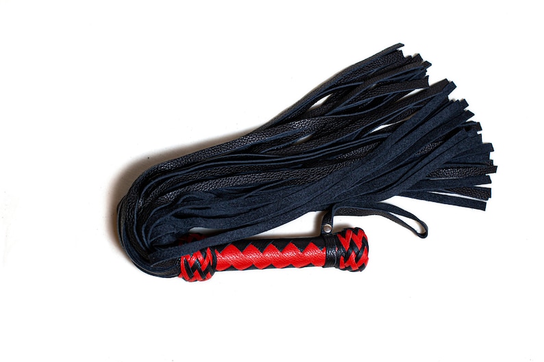 May include: A black leather flogger with a red and black handle. The flogger features multiple black leather falls and a handle with a red center and black braided ends. The item is set against a white background.