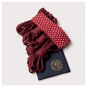 May include: A set of four coiled burgundy ropes bundled together with a red fabric band featuring white polka dots. A dark navy blue fabric pouch with a gold circular monogram logo containing the letters MDB is positioned beneath the ropes.