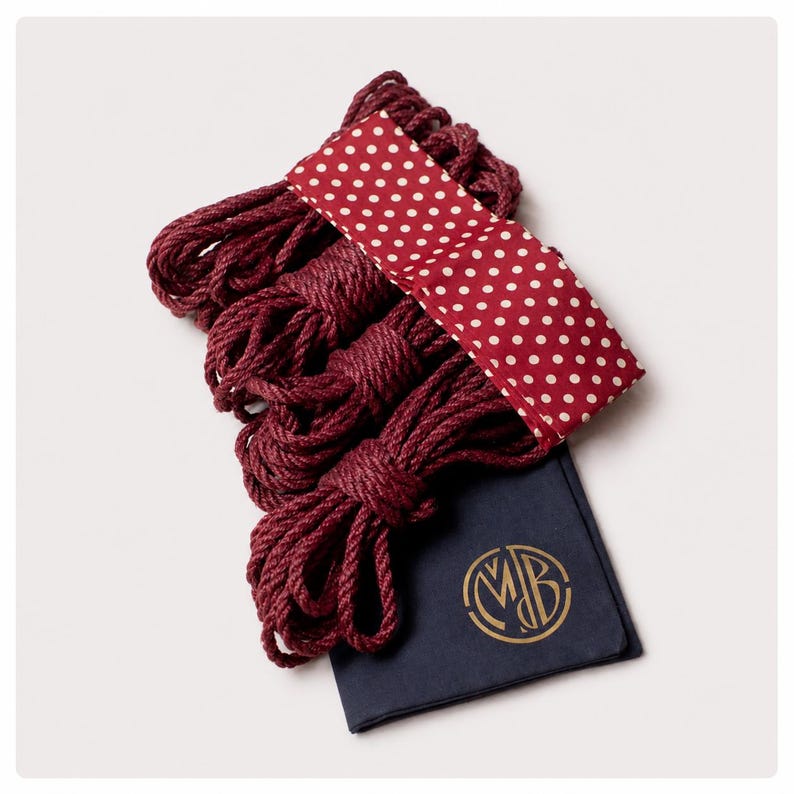 May include: A set of four coiled burgundy ropes bundled together with a red fabric band featuring white polka dots. A dark navy blue fabric pouch with a gold circular monogram logo containing the letters MDB is positioned beneath the ropes.
