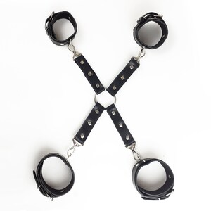 May include: A black restraint harness featuring four adjustable wrist or ankle cuffs connected by two intersecting straps. The straps are secured with silver-toned metal rivets and attached to a central metal ring using swivel snap hooks.