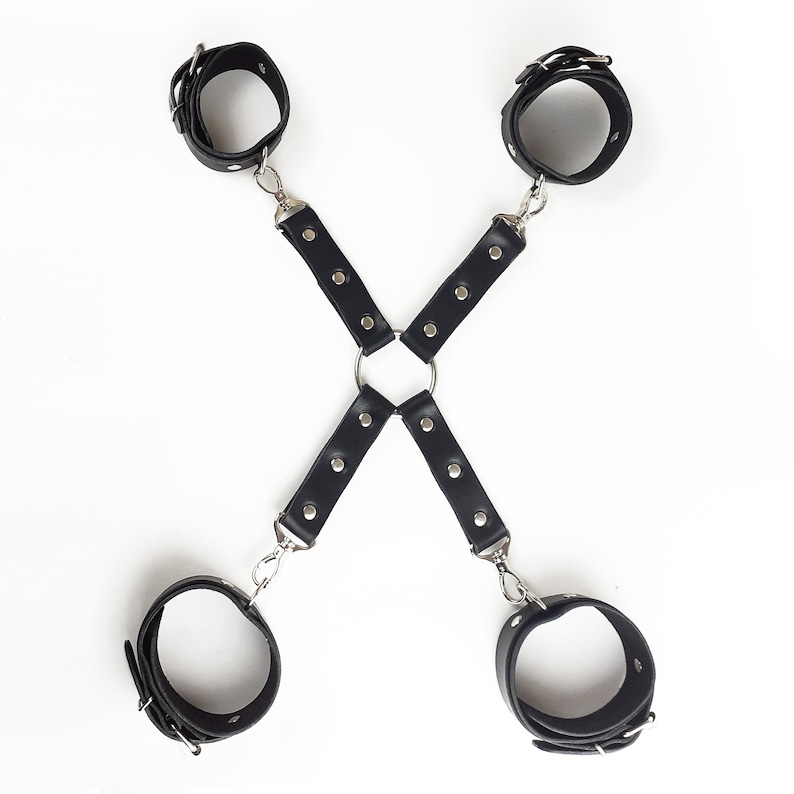 May include: A black restraint harness featuring four adjustable wrist or ankle cuffs connected by two intersecting straps. The straps are secured with silver-toned metal rivets and attached to a central metal ring using swivel snap hooks.
